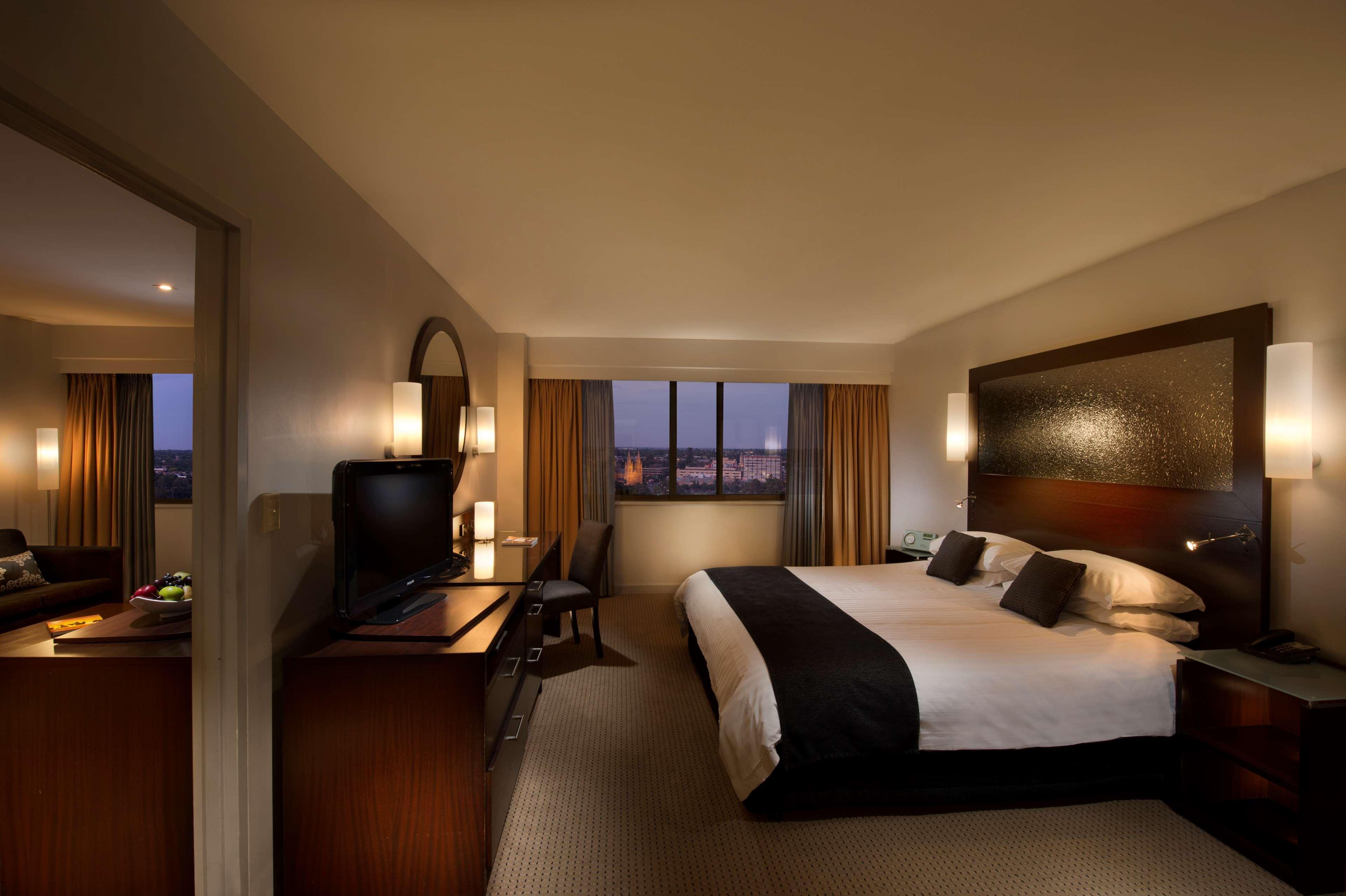 Stamford Plaza Adelaide Hotel Buitenkant foto The photo features a modern hotel room that includes a large bed with dark and light bedding, complemented by two pillows. The room is well-lit with warm lighting and has a contemporary aesthetic. To one side, there is a wooden dresser with a flat-sc