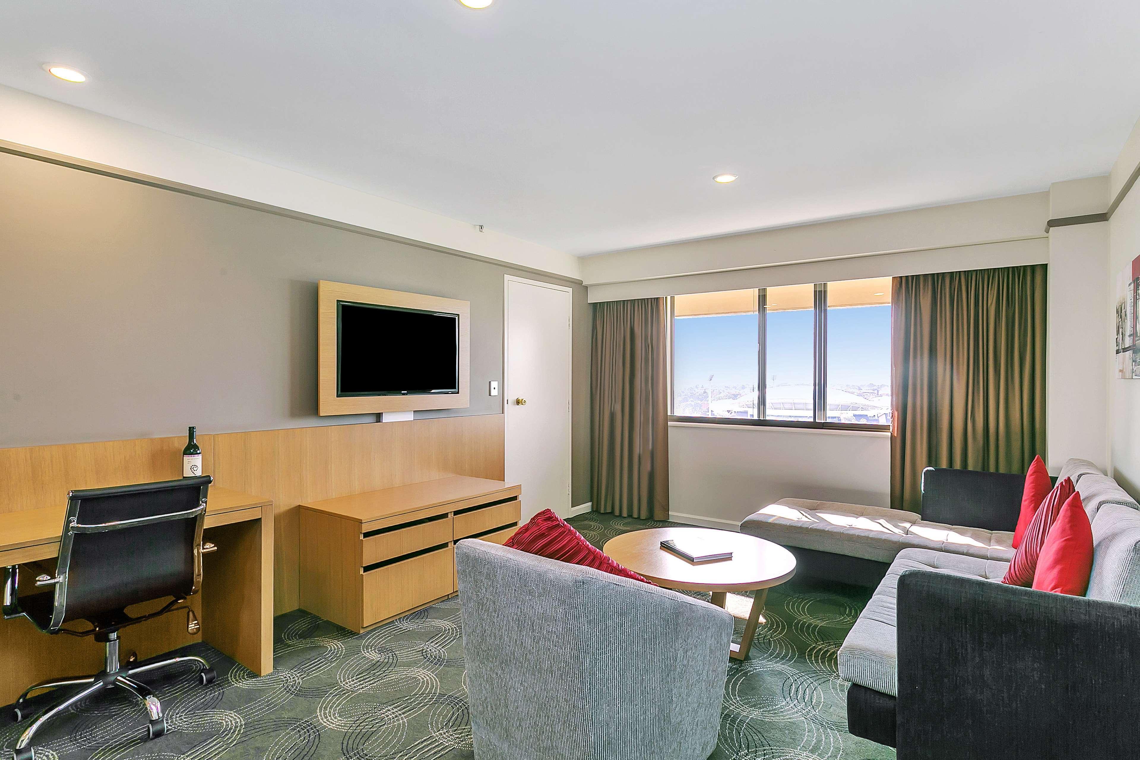 Stamford Plaza Adelaide Hotel Buitenkant foto The photo shows a modern hotel room or suite that features a cozy seating area. There is a comfortable sofa with red accent pillows, a round coffee table in the center, and a sleek desk with an office chair. On the wall, there is a flat-screen TV mou