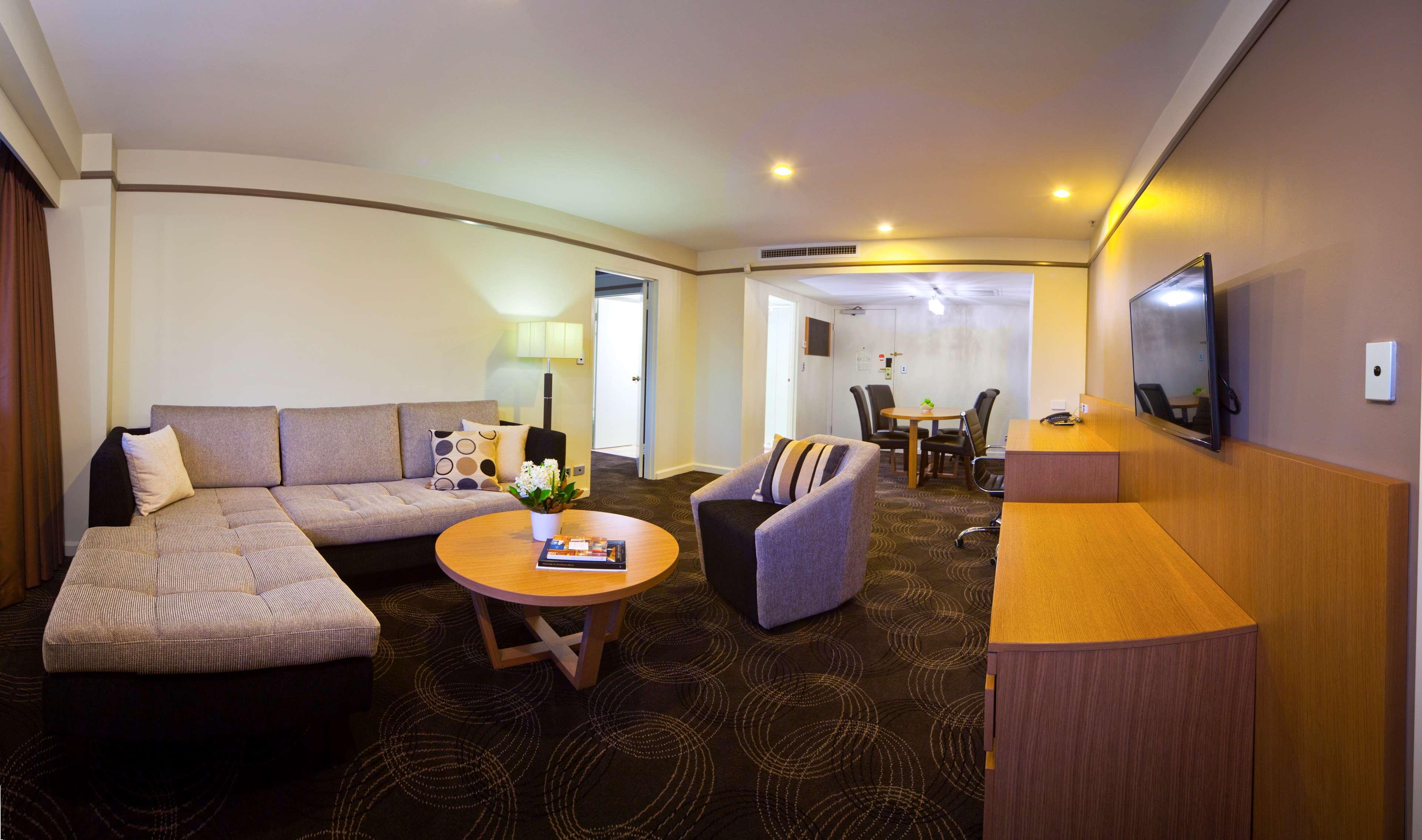 Stamford Plaza Adelaide Hotel Buitenkant foto The photo shows a spacious living area with modern decor. On the left, there is a light gray sectional sofa, accompanied by a round wooden coffee table in front of it. A couple of decorative pillows add a touch of color to the sofa. To the right, the