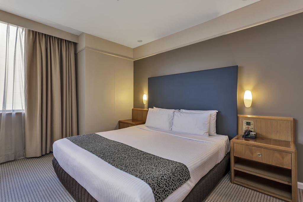 Stamford Plaza Adelaide Hotel Buitenkant foto The photo shows a modern hotel room. It features a double bed with a white duvet and a decorative grey and black throw. The headboard is a solid dark color. There are two bedside tables on either side of the bed, each with a lamp. Natural light enter